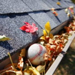 White Rain Gutter Full of Autumn Leaves. Baseball also in gutter. Grey shingle roof