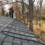 Heat cable zig zagged on composite shingle roof. Dark grey roof. Fall day. dreary weather. Man on roof is sweeping (possibly to remove leaves).