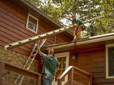 KGuard installation: one man on the ground is passing a gutter up to a man standing on the roof of a house. Ladder visible. house is a dark red or brown color. Gutter appears to be brown. You can see the brackets inside the gutter and the primed interior color.