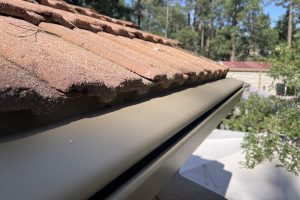 Cream K-Guard hooded gutter running along the edge of a tile roof on a Colorado home, with pines behind