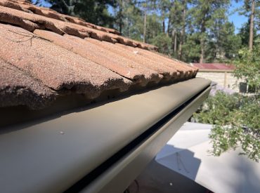 Cream K-Guard hooded gutter running along the edge of a tile roof on a Colorado home, with pines behind