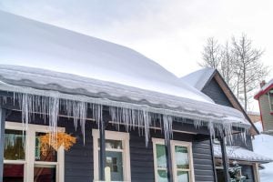 dark blue home covered in thick snow. Gutters full of ice. Icicles hanging. Ice dam. ice overflow. Frozen Downspouts. Water can't exit downspout as needed.