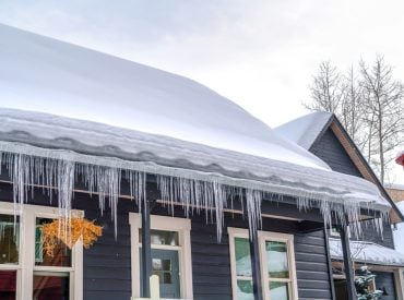 dark blue home covered in thick snow. Gutters full of ice. Icicles hanging. Ice dam. ice overflow. Frozen Downspouts. Water can't exit downspout as needed.